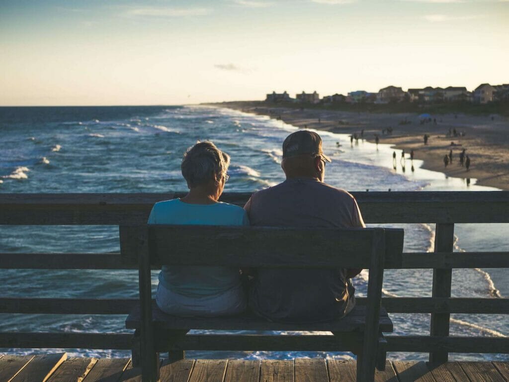 A couple sits on the bench of a pier, overlooking the coastline at sunset.