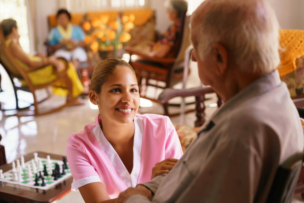 A young nurse kneels beside an older man and smiles at him. They are in a common room in a senior care facility.