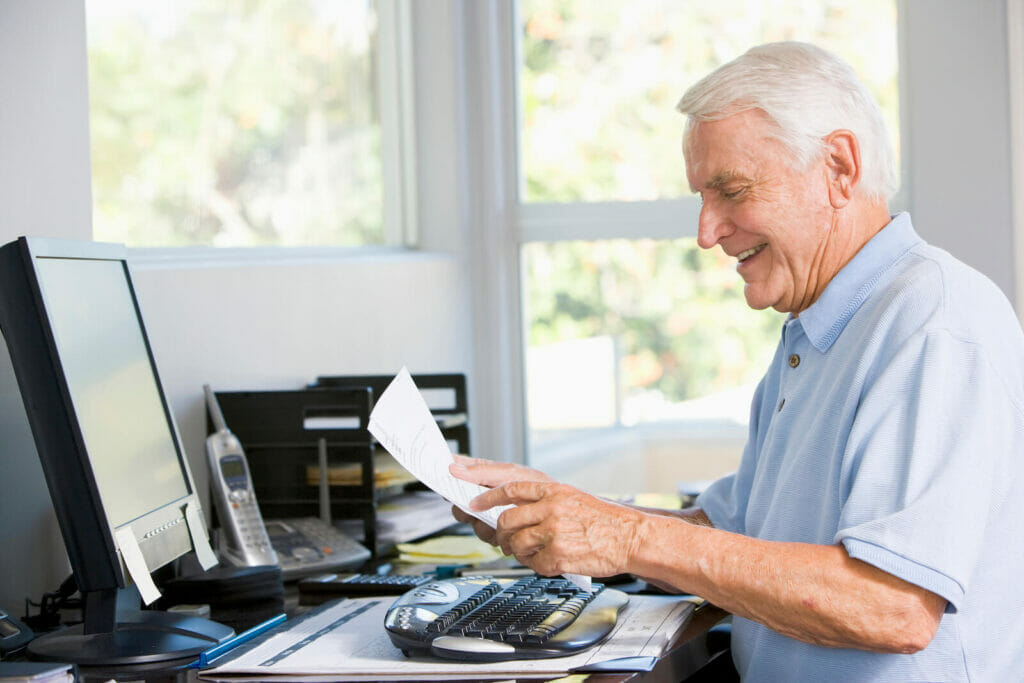 An older adult man looks at paperwork while sitting at a desk with a desktop computer.