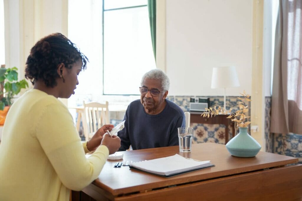 A woman and older man sit at a table in a home. The woman helps the man with paperwork.