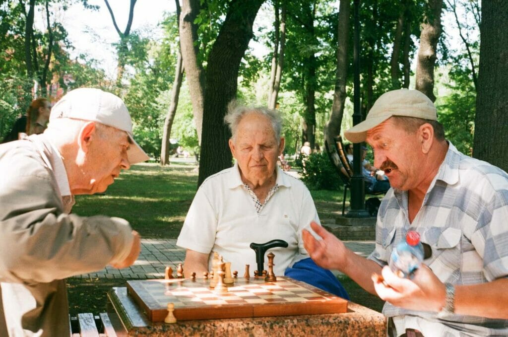 A group of three older adult men sit at a table outside and play chess.