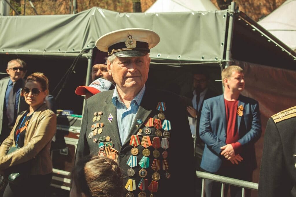 An older man stands outside in the sun. He wears a suit jacket decorated with medals from serving in the armed forces.