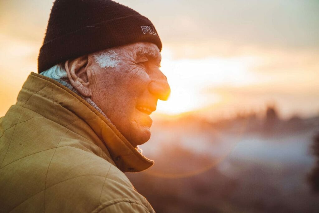 A side portrait of an older adult man wearing a hat. The sunrise is behind him.