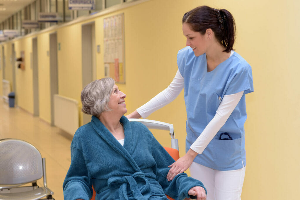 A youthful female nurse smiles at an older adult woman in a wheelchair. They are in a nursing home hallway.