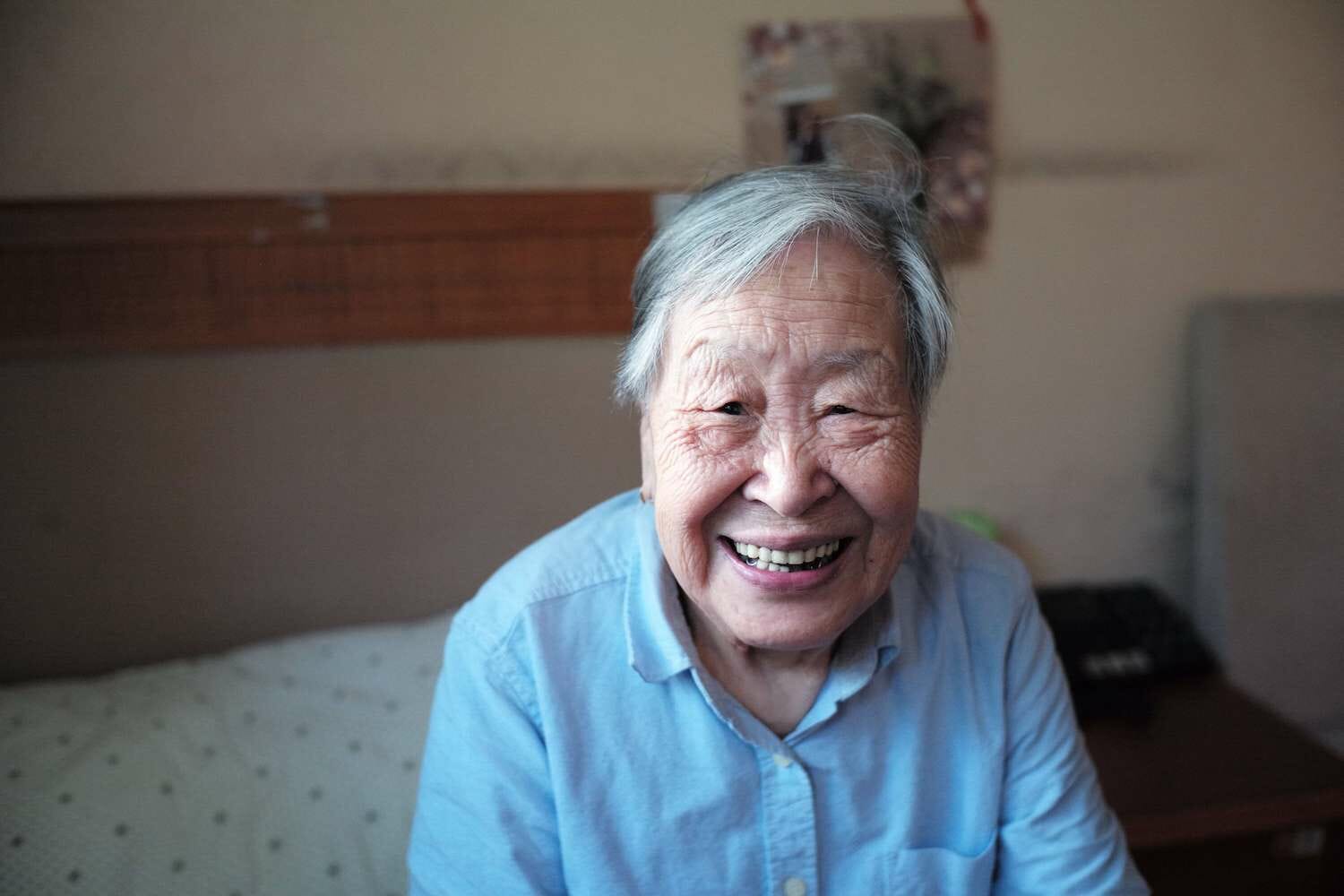 A portrait of an older adult woman sitting in a senior care facility room.