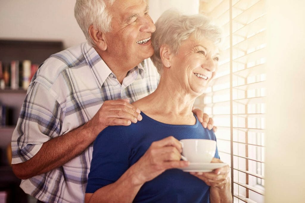 senior couple standing looking out a sunny window with the woman having a cup of coffee