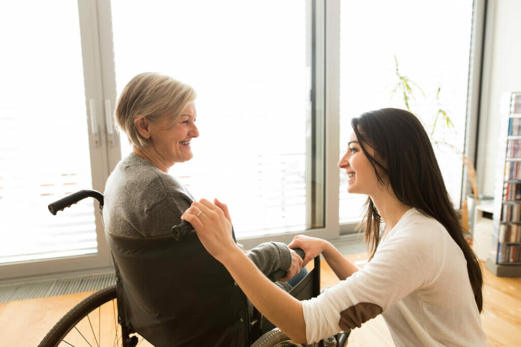 A young woman kneels beside an older woman who sits in a wheelchair. They are smiling at each other.